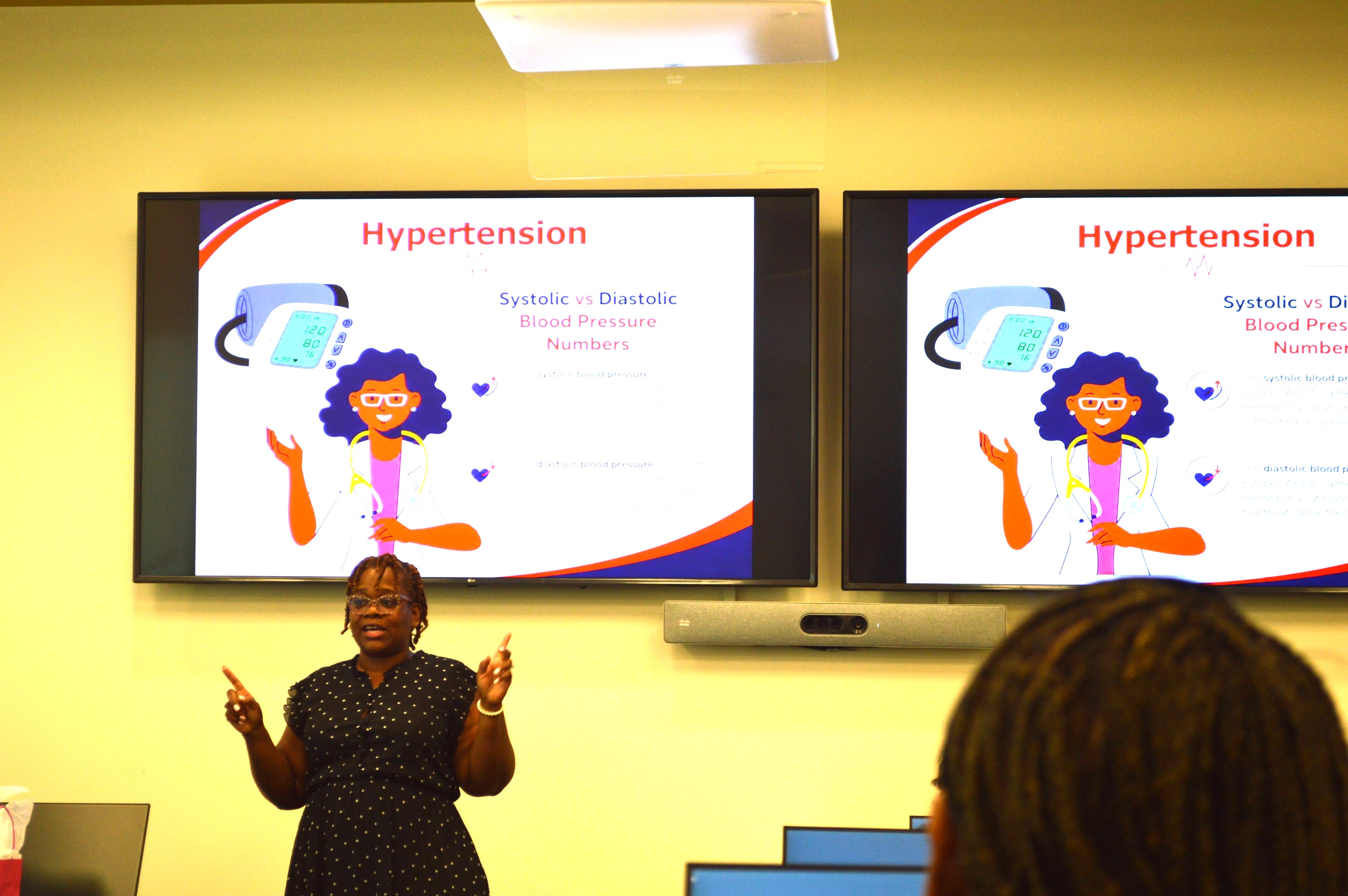 A woman points at video screens with information about hypertension displayed on them.